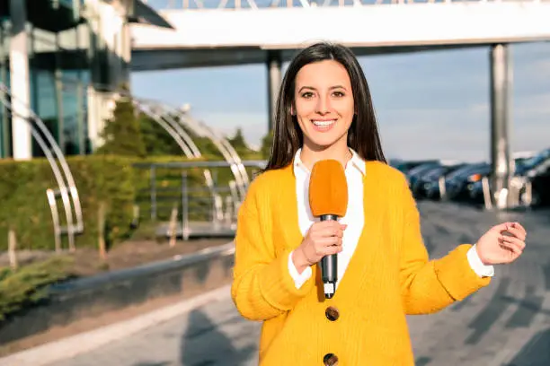 Photo of Young female journalist with microphone working on city street