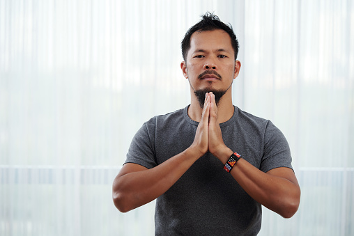 Serious religious young man making namaste gesture and looking at camera