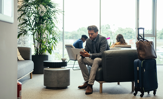 Handsome business man sitting in armchair with carry on luggage and working on digital tablet while waiting for flight at airport lobby.