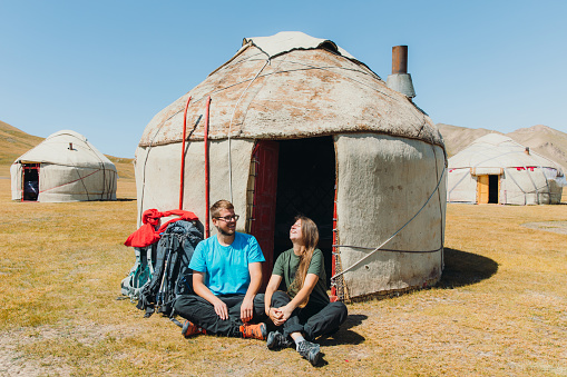 Happy couple with backpacks sitting at the meadow near the traditional yurt enjoying the sunny summer morning at Tian Shan mountains, Central Asia