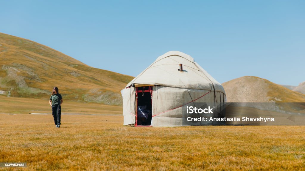 Young smiling woman meets morning in the mountains from yurt camp in Kyrgyzstan Woman explorer with long hair walking at the meadow near the traditional yurt enjoying the sunny summer morning at Tian Shan mountains, Central Asia Bishkek Stock Photo