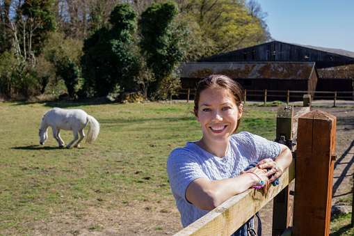 A mid adult woman standing and leaning on a wooden gate while standing in a field on her home farm. Her pet horse is standing in the background, grazing in the field. The woman is looking at the camera and smiling.