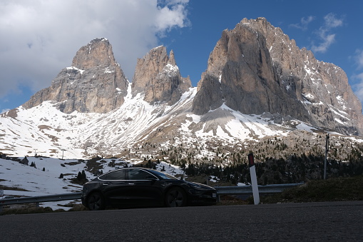 Selva di Val Gardena, Bolzano, Italy - June 3, 2021: A static shot of a solid black Tesla Model 3 dual motor with tinted windows and aero wheels. Snow-covered mountain; sunny spring day. Passo Sella