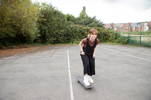 Young woman wearing dungarees and brown t-shirt, with a skateboard in an abandoned tennis court in the city of Nottingham in the midlands of England