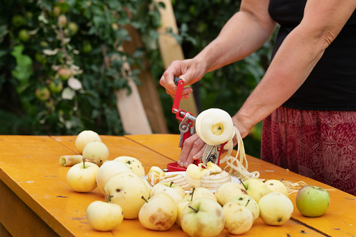 Woman peels apples just picked up from the tree using an apple peeler device on the orange wooden table in the garden