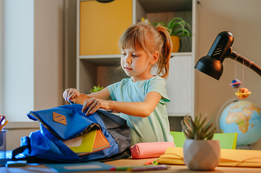 Elementary female student packing backpack for school at home. Selective focus.