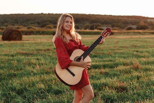 Young blonde plays guitar and smiles in a field at sunset