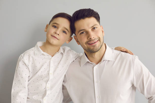 istock Father with son who are dressed in white shirts and cuddling while standing on gray background. 1339481727