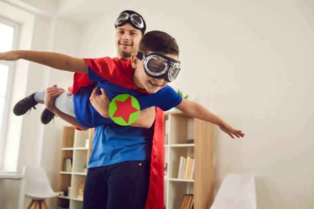 Father helps his son fly like a superhero. Boy with pilot glasses play fly with his dad at home. Dream big. Loving father helps his son fly like a superhero. Boy with pilot glasses play fly with his dad at home. Cheerful family in red cloaks and pilot glasses are having fun together. happy fathers day funny stock pictures, royalty-free photos & images