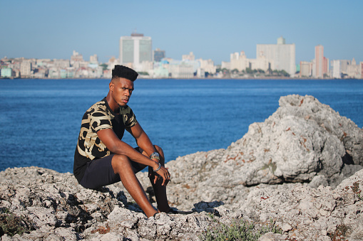 Portrait of young black Cuban dressed in a shirt and black pants. The young man poses on the coast of the bay of Havana and looks out to sea. Caribbean beauty.