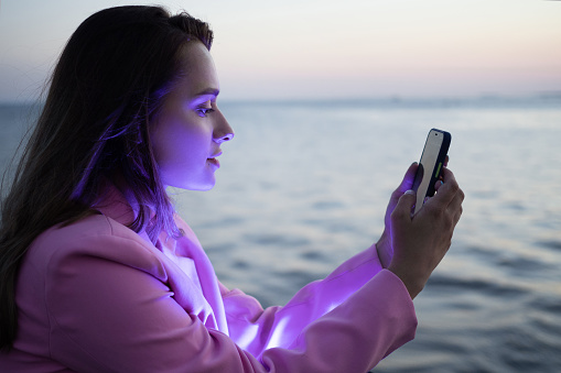 A beautiful laughing brunette uses a smartphone to communicate, a young woman sits against the background of the sea in the evening with a smartphone in her hands, a bluish glow on her face