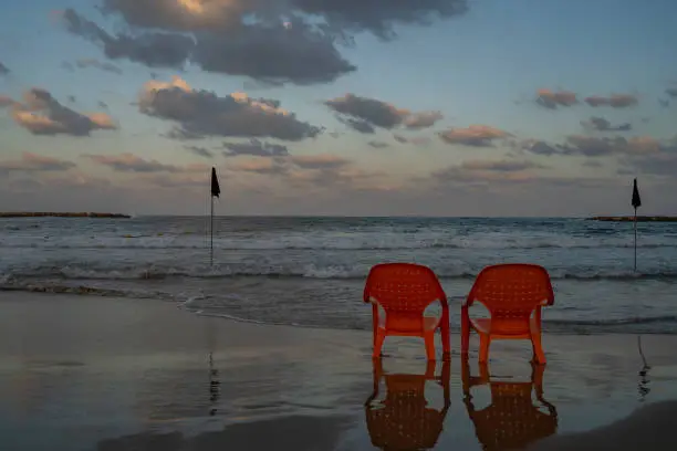Two empty, orange colored beach chairs, standing by a rough sea with black flags at dawn.
