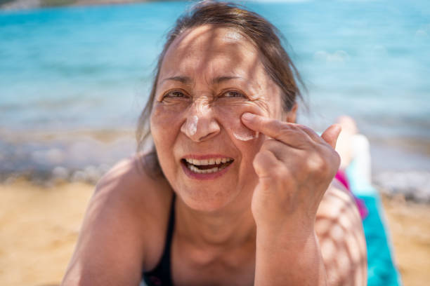 femme âgée appliquant un écran solaire sur son visage et souriant tout en prenant un bain de soleil sur la plage - outdoor chair furniture travel vacations photos et images de collection