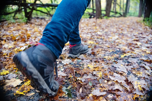 Soft focus, low angle photograph of an unrecognizable man wearing hiking sneakers and walking on a trail covered with autumn leaves.