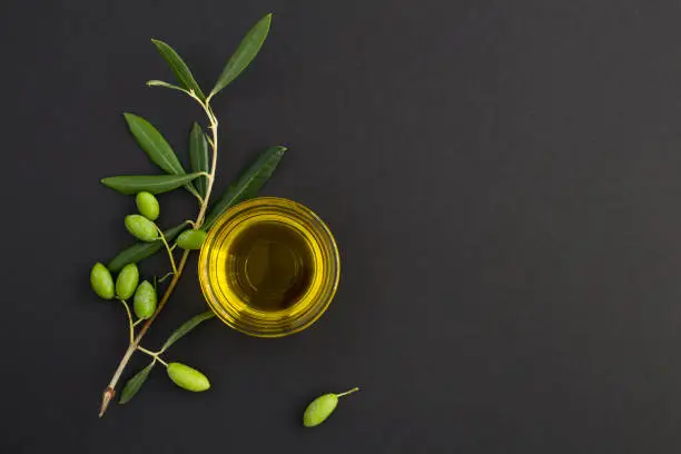Top view of olive oil in a glass bowl and branch with green olives on the black background. Copy space.