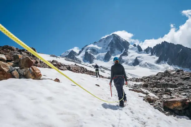 Two females Rope team members on acclimatization day dressed in mountaineering clothes walking in crampons with ice axes by snowy slopes in a climbing harness and dynamic rope on close-up foreground