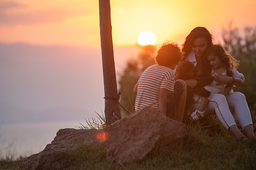 Portrait of happy little girl, boy and young woman having good time together with pet dog during sunset in outdoor. The sky is orange due to sunset. Sun is seen in frame. Shot with a full frame mirrorless camera.
