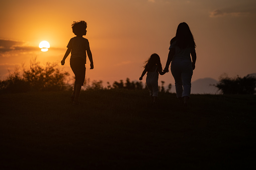 Silhouette of mother, daughter and son walking toward sun during sunset in outdoor. The background is orange colored sky. They are seen in full length. Shot with a full frame mirrorless camera.