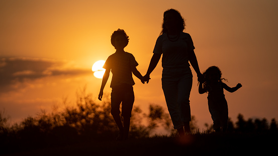 Silhouette of mother, daughter and son walking toward sun during sunset in outdoor. The background is orange colored sky. They are seen in full length. Shot with a full frame mirrorless camera.