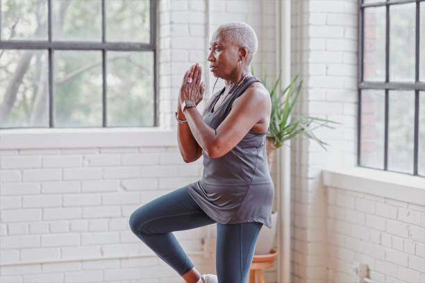 Active senior woman doing yoga at home A retired black woman does yoga at home in her modern loft apartment to stay active and healthy. She is holding a standing balancing pose. balance stock pictures, royalty-free photos & images