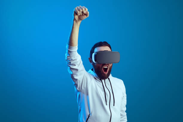 istock Man wearing VR headset on, standing with raised hand and screaming over blue 1339528900