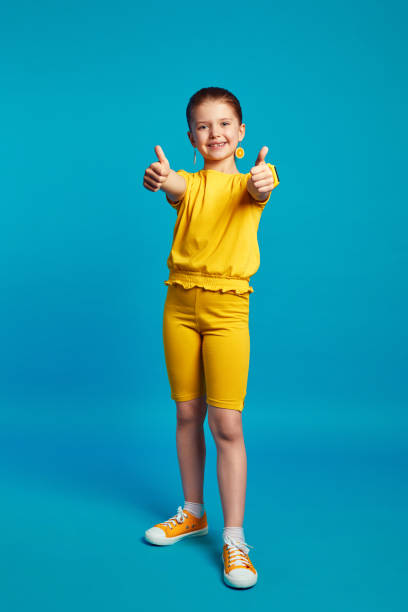urocza dziewczynka w żółtym stroju uśmiechająca się i pokazująca kciuki w górę na tle niebieskiego - endorsement appreciate validate thumbs up zdjęcia i obrazy z banku zdjęć
