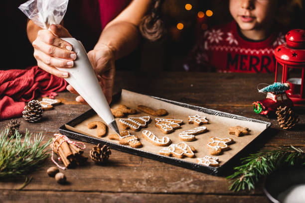 Woman icing gingerbread christmas cookie by son in kitchen Woman decorating gingerbread cookie with icing from bag. Boy is standing by mother making Christmas sweets. They are in kitchen. baking stock pictures, royalty-free photos & images