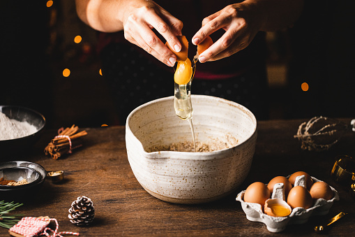 Hands breaking brown eggshells over bowl. Egg yolk is falling in container. Woman is preparing Christmas cookies.