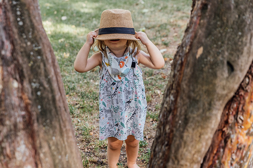 Funny little girl wearing a colorful dress covering her face with her hat in the park. View through two trees of a little girl standing covering her face with her hat.