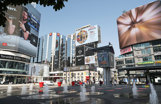 Toronto, Canada - August 26, 2021: Colorful signs dominate Yonge-Dundas Square downtown. Small fountains spurt water around the popular outdoor venue on a summer morning.\n1 Dundas Street East.
