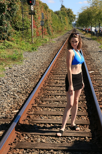 A model posing between the tracks of a railroad. She is wearing black sandals and mini dress and a blue bikini top.