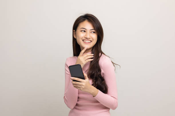 istock Pretty young asian woman using smartphone standing on isolated white background feeling happy. Shopping online payment with mobile phone. Excited and surprised female holding cellphone. 1339577598