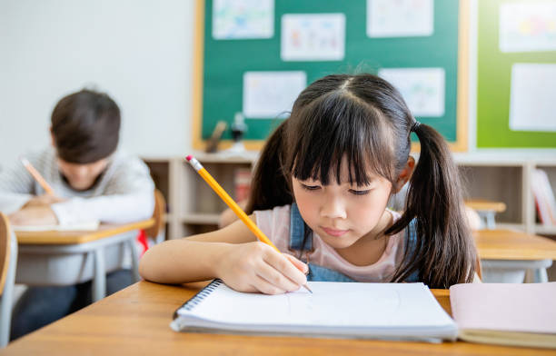 ritratto di un piccolo alunno che scrive alla scrivania in classe alla scuola elementare. studentessa che fa il test nella scuola primaria. bambini che scrivono appunti in classe. concetto di conoscenza dell'istruzione - korea child baby asian culture foto e immagini stock