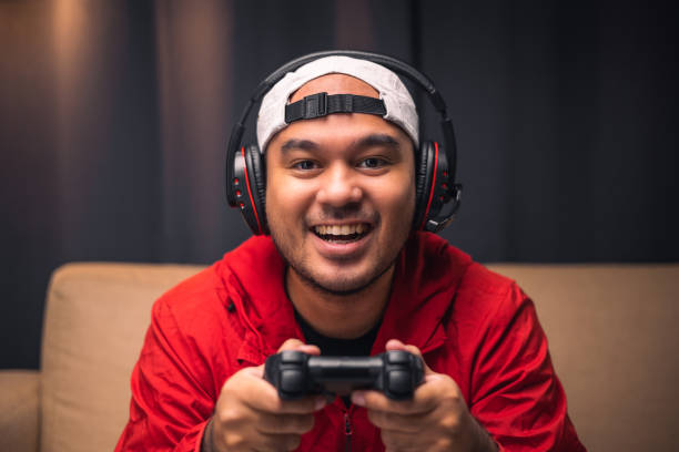 istock Playing video game. Young asian handsome man sitting on sofa holding joystick in living room. Happiness Streamer Indian man wearing headset playing game online in the darkroom. 1339579727