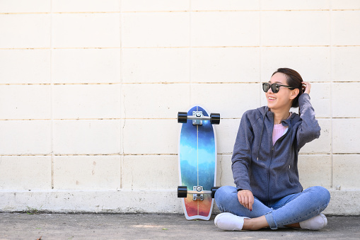 Happy Asian woman wearing sunglasses with surfskate skateboard sitting against concrete wall