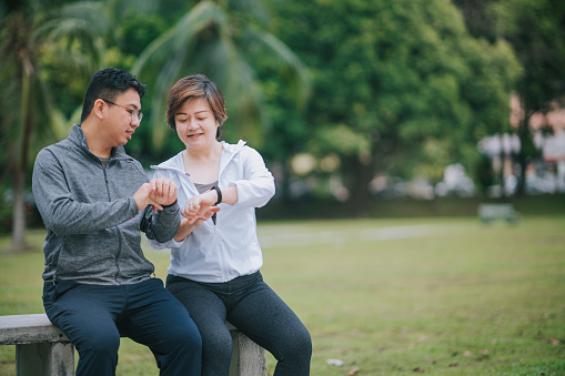 asian chinese mid adult couple using fitness tracker mobile app connecting to fitness tracker at public park after exercise in the morning at park bench
