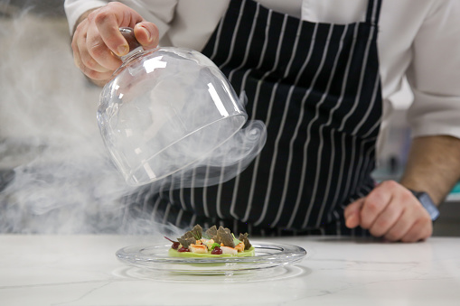 La mano del chef levanta el cloche de vidrio de un plato con comida caliente y humo en movimiento en el restaurante. Plato exquisito, concepto creativo de comida de restaurante, comida de alta costura. photo
