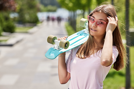 young beautiful brunette skateboarder girl in sunglasses with skateboard standing outdoors in park