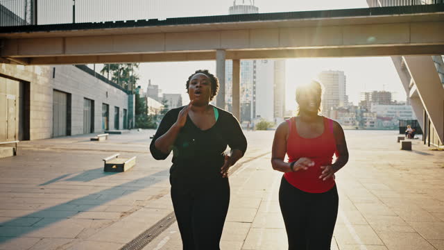 Two active overweight african american ladies practicing cardio workout, running together in city area in evening