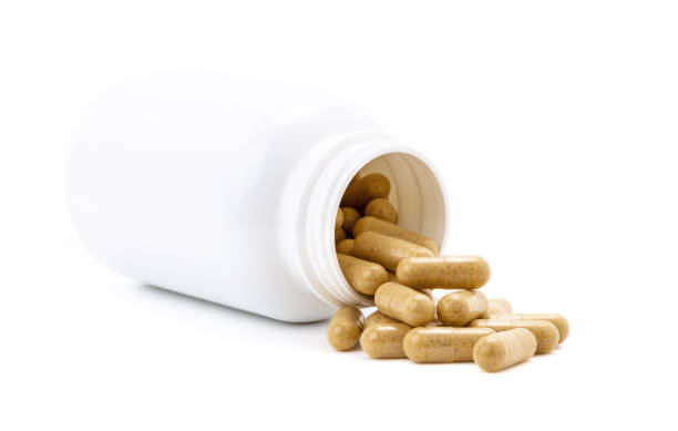 Brown herbal supplement capsules pour out from white opened plastic bottle