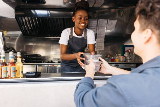 jeune femme en tablier travaillant au food truck. propriétaire d’une petite entreprise donnant des aliments emballés à emporter au client. - food truck photos et images de collection