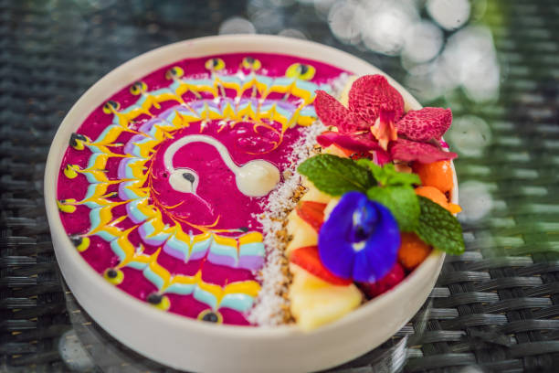 Healthy tropical breakfast, smoothie bowl with tropical fruits, decorated with a pattern of colorful yogurt with turmeric and spirulina. It is also decorated with fruits, flowers, chia seeds, coconut, granola, pineapple, mint and strawberries
