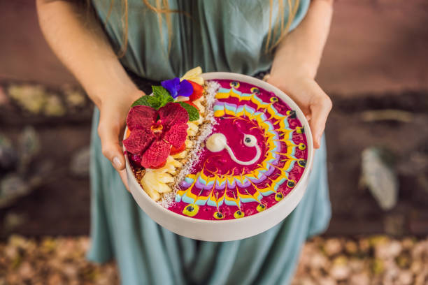 Young woman having a mediterranean breakfast, eats Healthy tropical breakfast, smoothie bowl with tropical fruits, decorated with a pattern of colorful yogurt with turmeric and spirulina. It is also decorated with fruits, flowers, chia seeds, coconut
