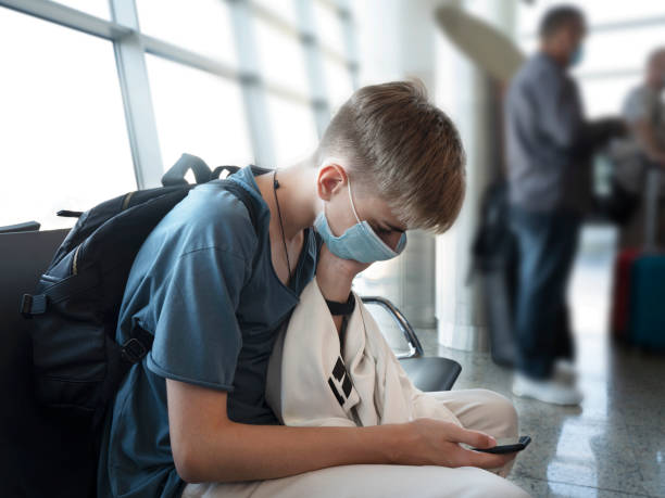 jeune homme sérieux adolescent de 15 à 18 ans tapant un message au téléphone alors qu’il était assis dans le salon de l’aéroport. jeune étudiant en tenue décontractée et masque de protection à l’aide d’un smartphone. - 12 13 years 13 14 years learning education photos et images de collection