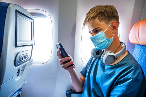 jeune adolescent sérieux portant un masque utilisant un téléphone dans un avion volant pendant la pandémie de covid-19. beau gars 15-18 ans échangeant des messages sur un téléphone portable tout en voyageant en avion - 12 13 years 13 14 years learning education photos et images de collection