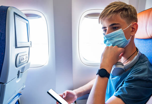 jeune adolescent sérieux portant un masque utilisant un téléphone dans un avion volant pendant la pandémie de covid-19. beau gars 15-18 ans échangeant des messages sur un téléphone portable tout en voyageant en avion - 12 13 years 13 14 years learning education photos et images de collection