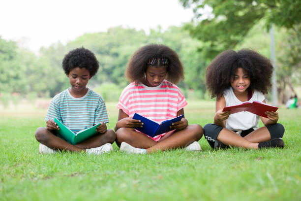 Children reading book on grass outdoor. Group of African American children in casual wear reading book while sitting on green grass in the park, against green summer garden Children reading book on grass outdoor. Group of African American children in casual wear reading book while sitting on green grass in the park, against green summer garden 9 stock pictures, royalty-free photos & images