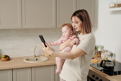 Young female with adorable baby daughter talking in video chat in the kitchen