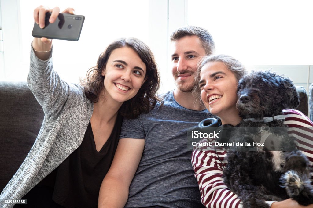 Group of  friends hugging their affectionate dog take a selfie taken with phone in a bright room. Group of  friends hugging their affectionate dog take a selfie taken with phone in a bright room.  Selfie of group of friends with black poodle. Pets Stock Photo
