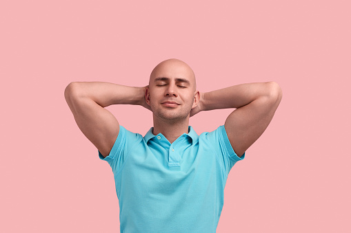 Calm pleased bald homosexual man with bristle is relaxing, closes eyes, keeps hands under head, lays on the ground, enjoys sun, gay friendly, wears blue polo shirt, poses over pink background